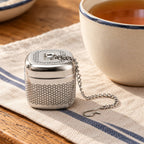 Square stainless steel tea infuser and chain beside a mug of amber tea