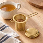 Gold mesh infuser prepared with herbal tea beside a ceramic cup
